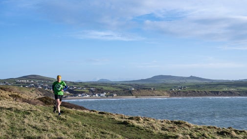 A runner wearing a green top and black shorts with a view down and across Aberdaron during a Trust10 run